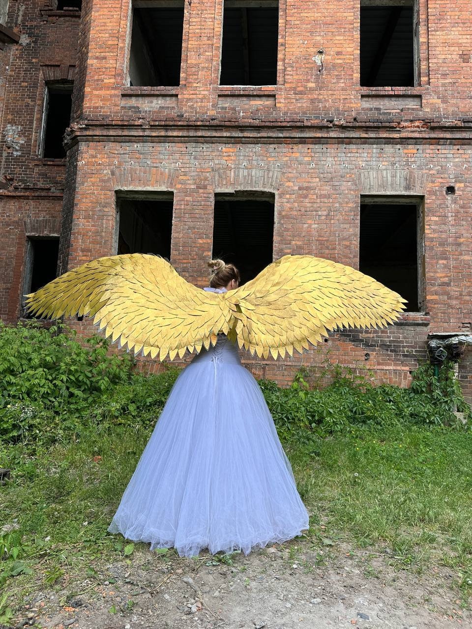 May include: A person wearing a white tulle dress and large gold angel wings, standing in front of a brick building.