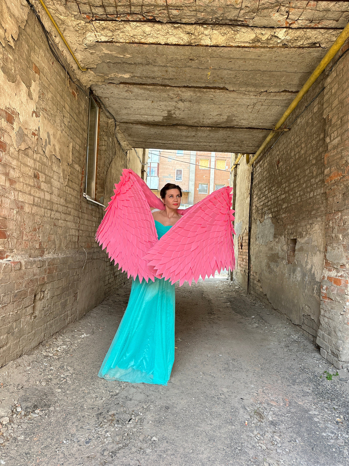 May include: A woman wearing a turquoise dress and large pink angel wings stands in a narrow brick alleyway.