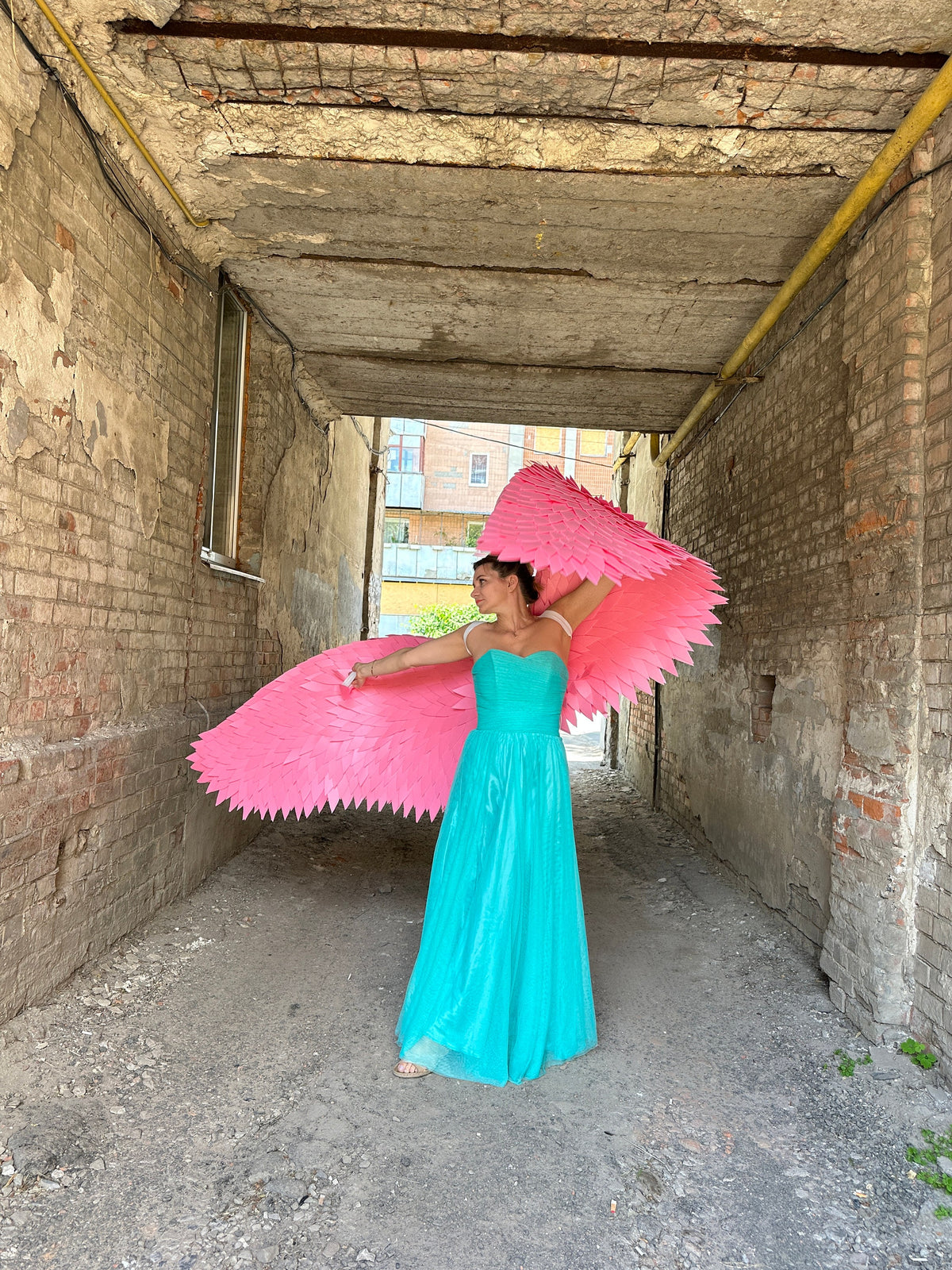 May include: A woman wearing a turquoise gown and pink angel wings stands in a narrow alleyway.