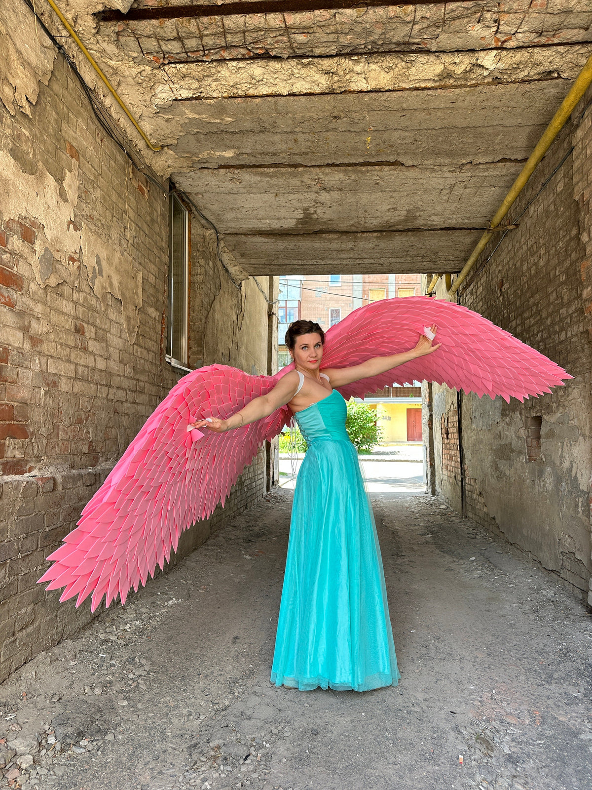 May include: A woman wearing a light blue dress with large pink angel wings. The wings are made of feathers and are spread out behind her. She is standing in a brick alleyway.