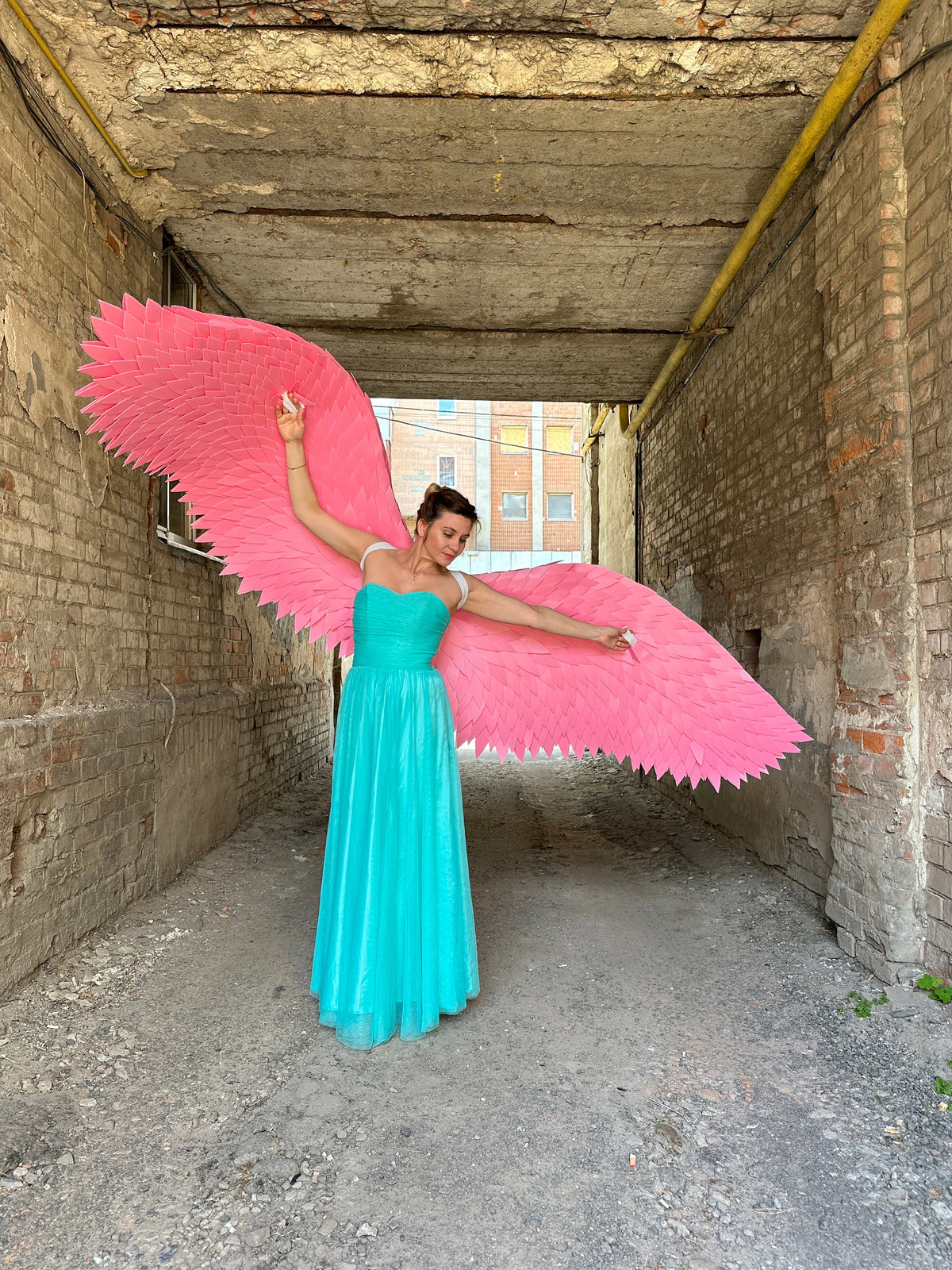 May include: A woman wearing a turquoise dress and large pink angel wings stands in a brick alleyway.