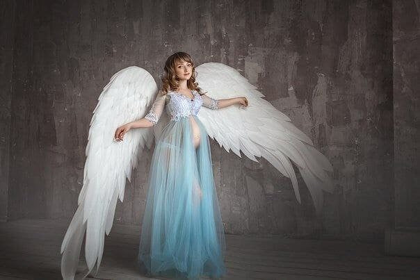 May include: A woman wearing a light blue sheer gown with white lace trim and large white angel wings. She is standing in front of a gray textured background.