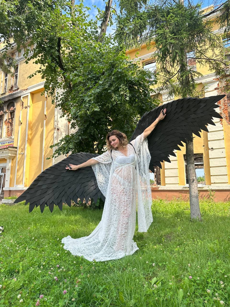 May include: A woman wearing a white lace dress and large black feathered wings stands in a grassy area. The wings are spread wide, and the woman is smiling. A yellow building with visible damage is in the background.