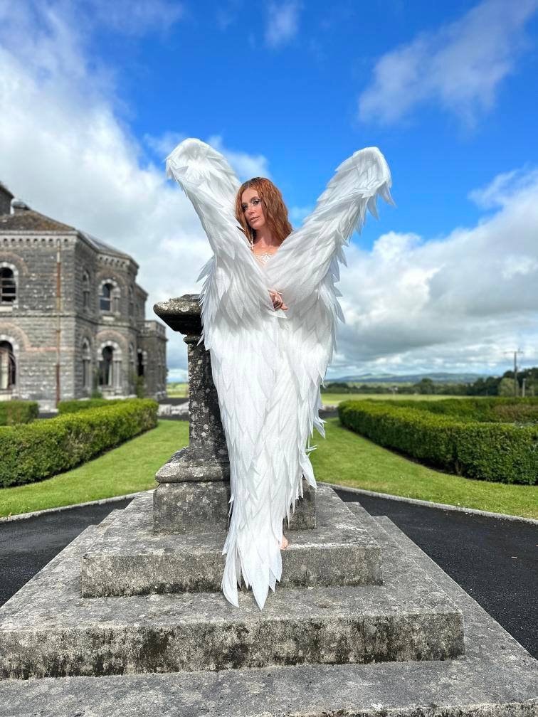 May include: A person wearing large, white feathered angel wings stands on a stone structure. The wings are spread wide against a backdrop of a blue sky with clouds, a large building, and green landscaping.