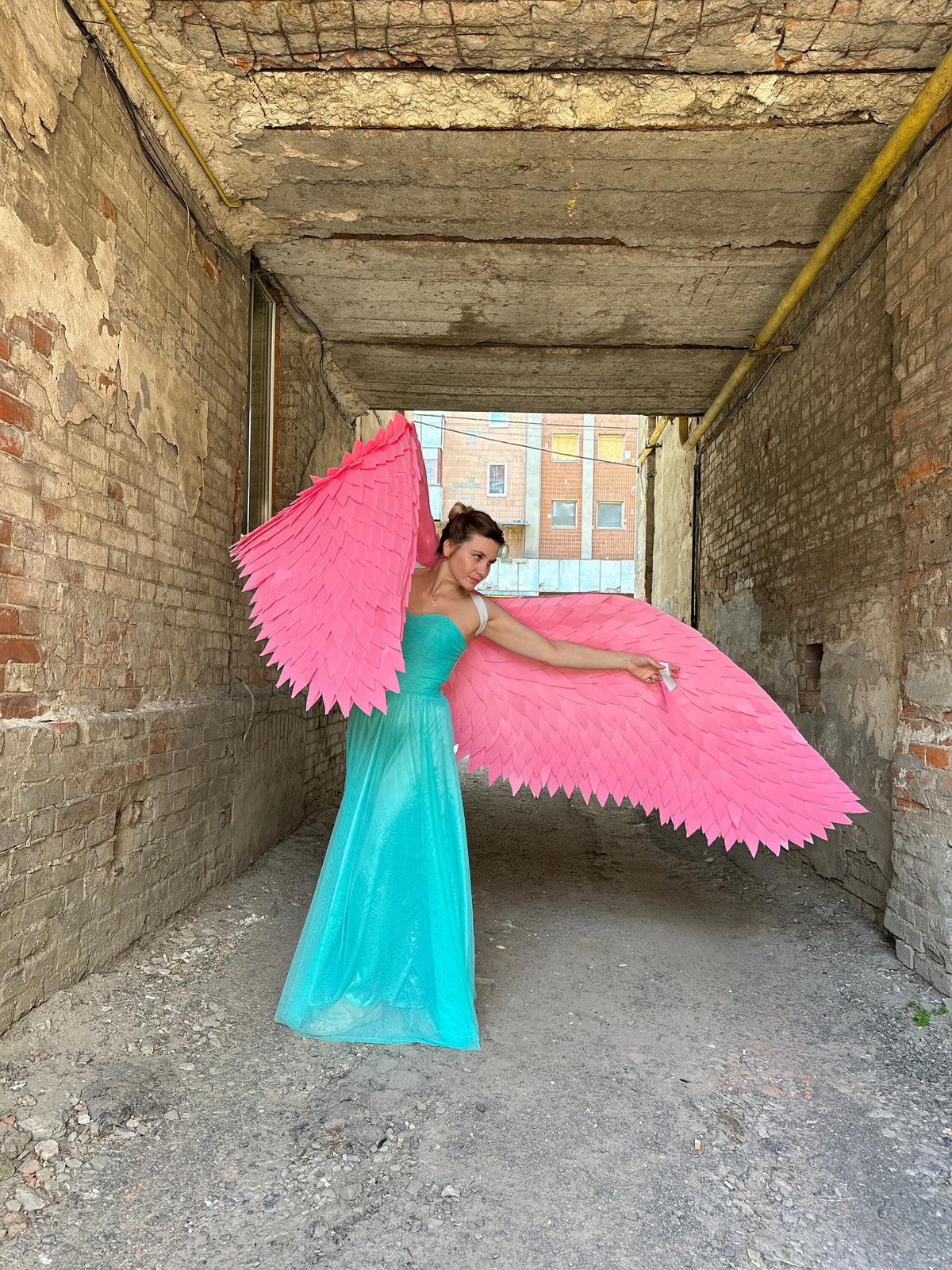 May include: A person wearing a turquoise gown with large pink wings in a brick alleyway.