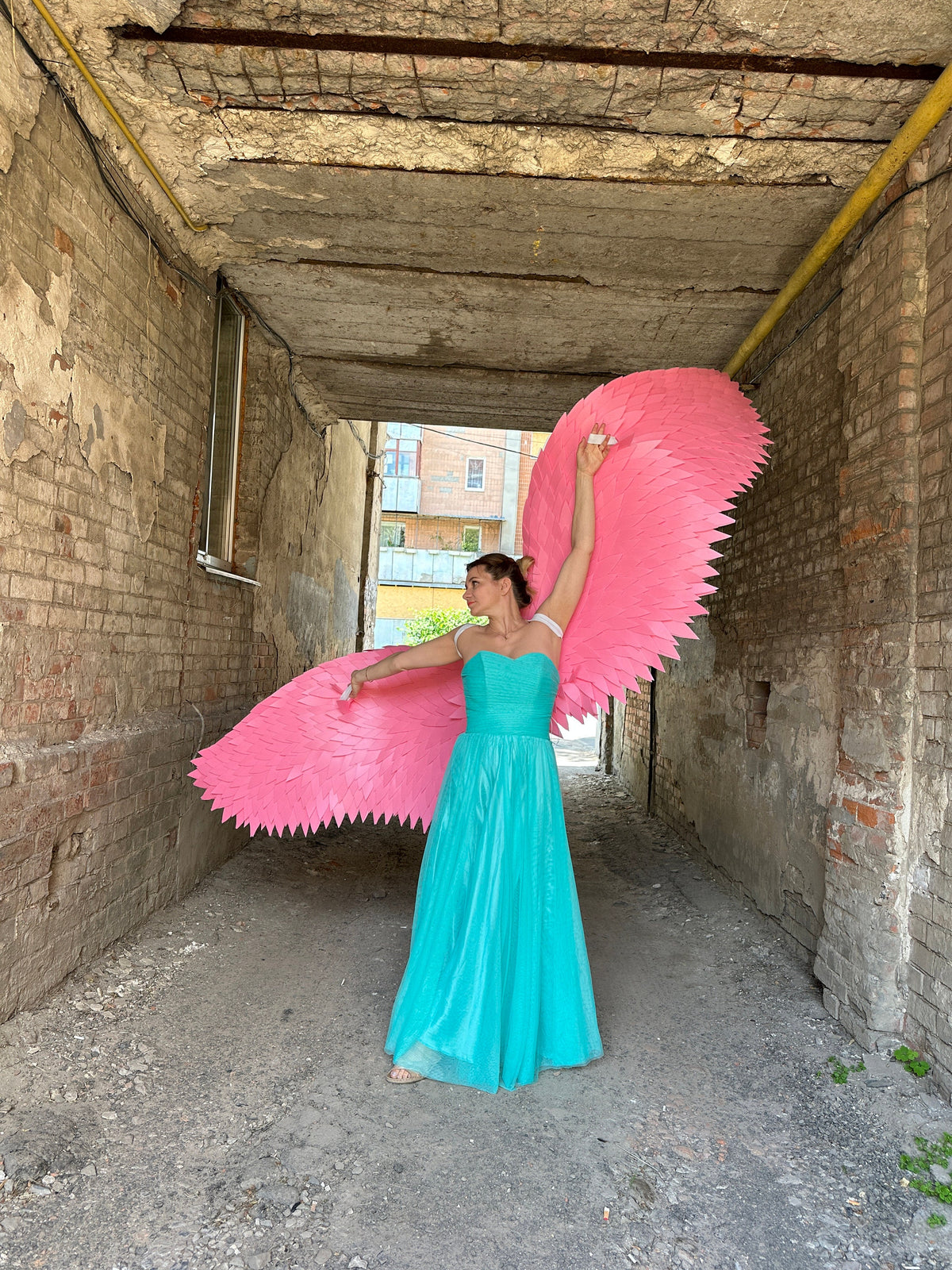 May include: A woman wearing a turquoise gown with large pink angel wings in a brick alleyway.