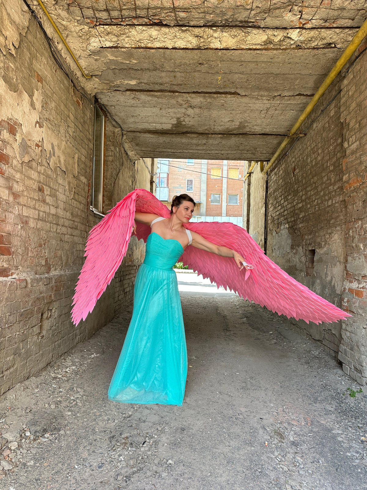 May include: A person wearing a turquoise dress and pink angel wings poses in a brick alleyway.