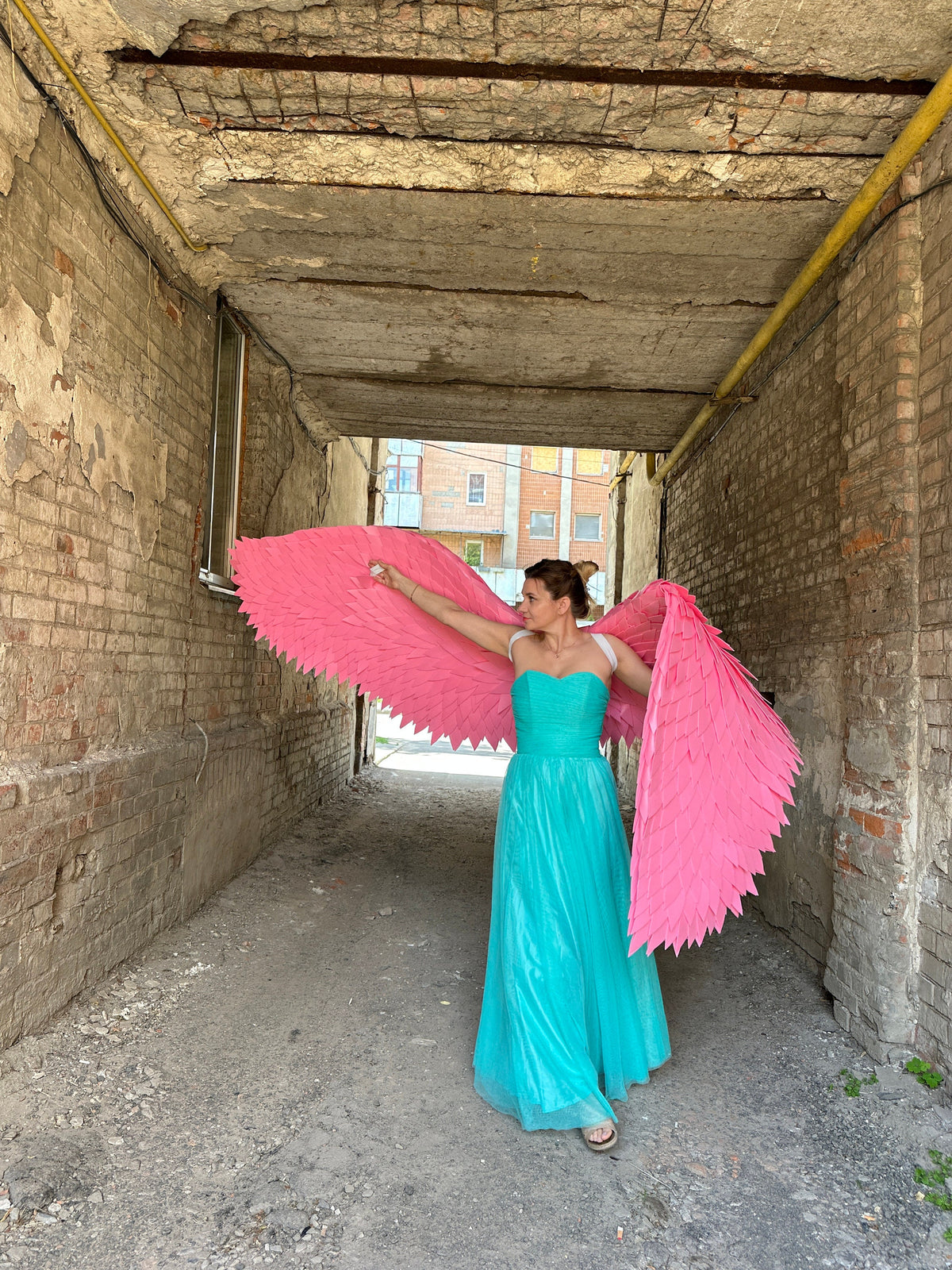 May include: A woman wearing a turquoise dress and large pink angel wings stands in a brick alleyway.