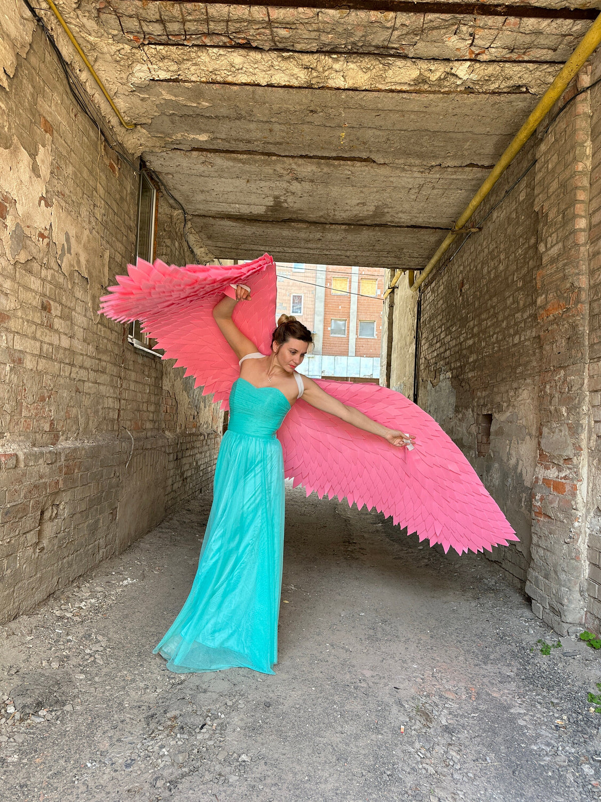 May include: A woman wearing a turquoise gown with pink angel wings in a brick alleyway.