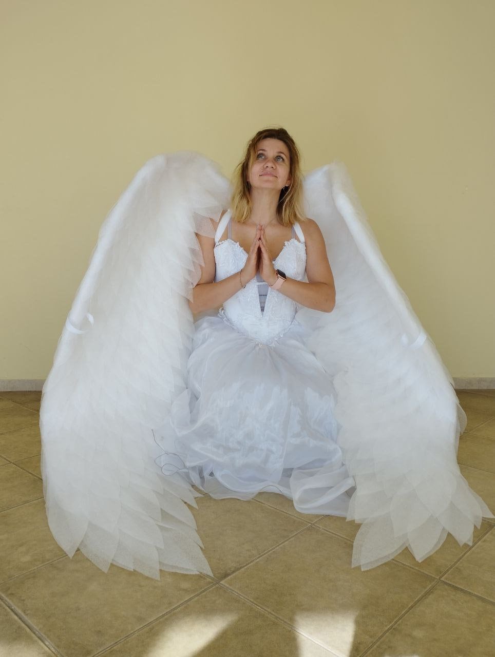 May include: A woman wearing a white dress and large white angel wings is kneeling with her hands clasped in prayer. She is looking up at the ceiling.