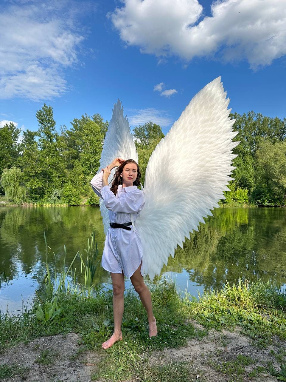 May include: A person wearing a white shirt and a black belt stands in front of a pond with large white feathered angel wings. The person is standing on a grassy bank with a blue sky and white clouds in the background.