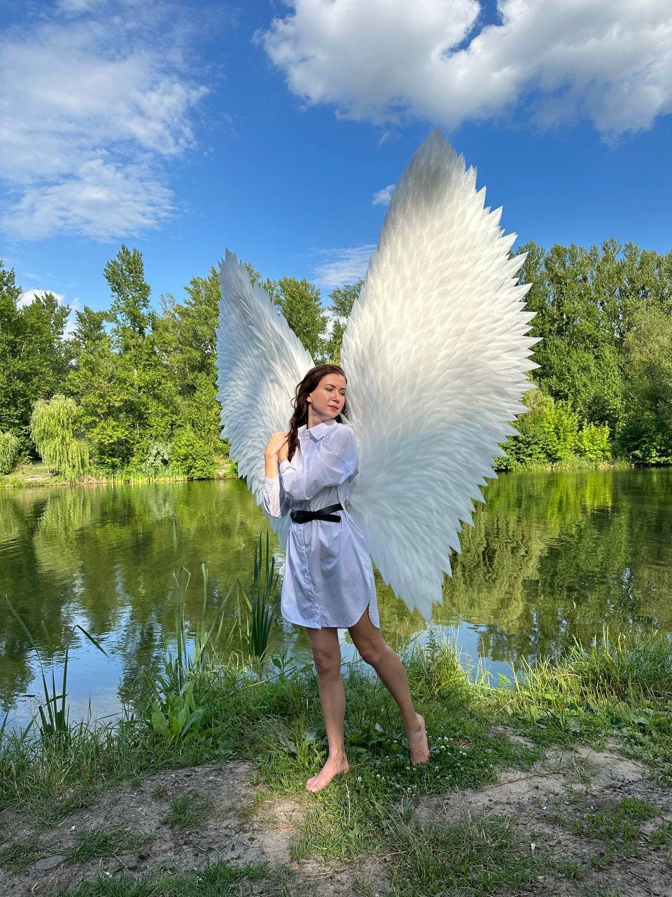 May include: A person wearing a white shirt and a black belt stands in front of a pond with large white angel wings behind them.