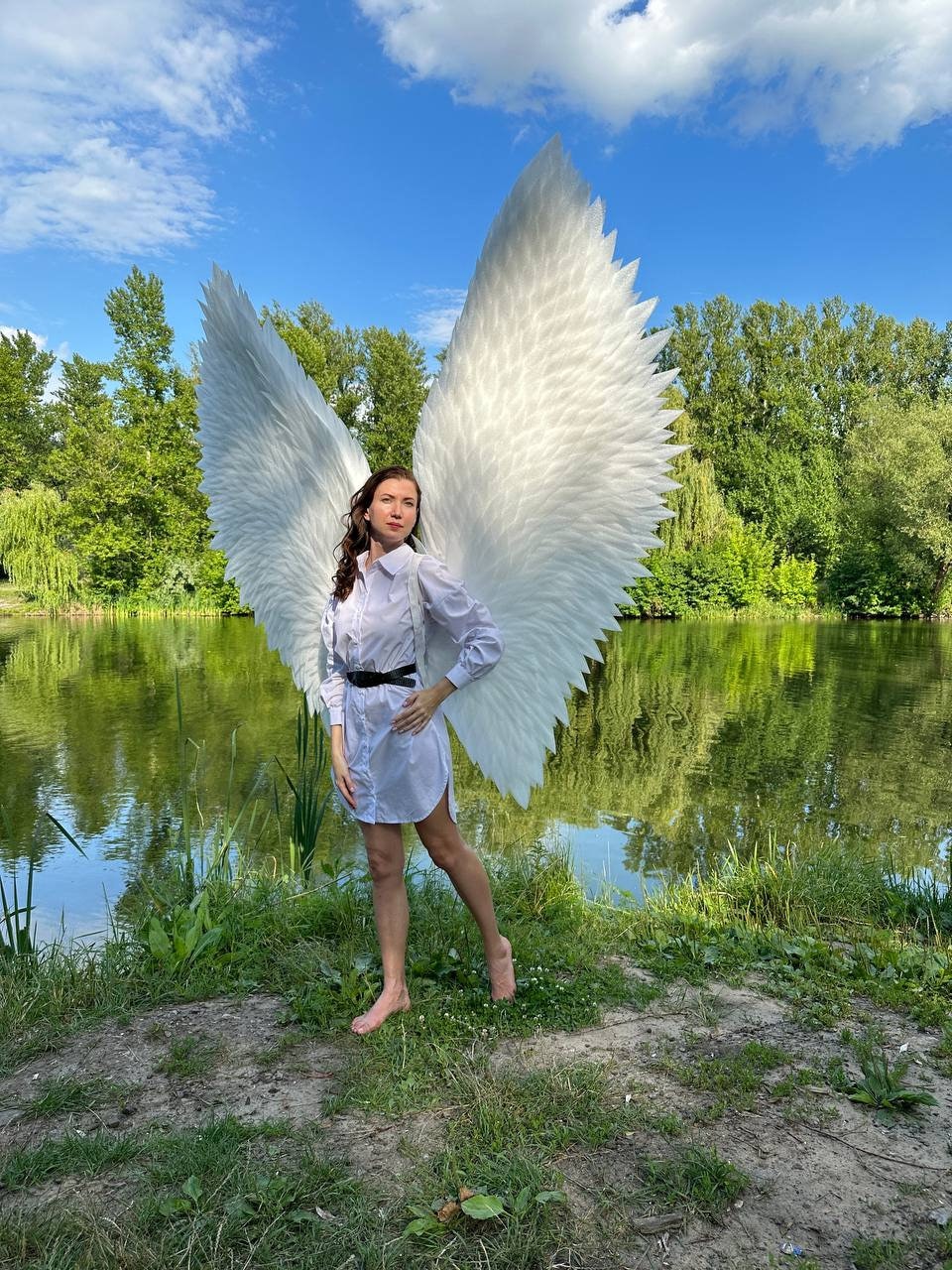 May include: A person wearing a white shirt and a pair of large white angel wings, standing in a grassy area by a pond.