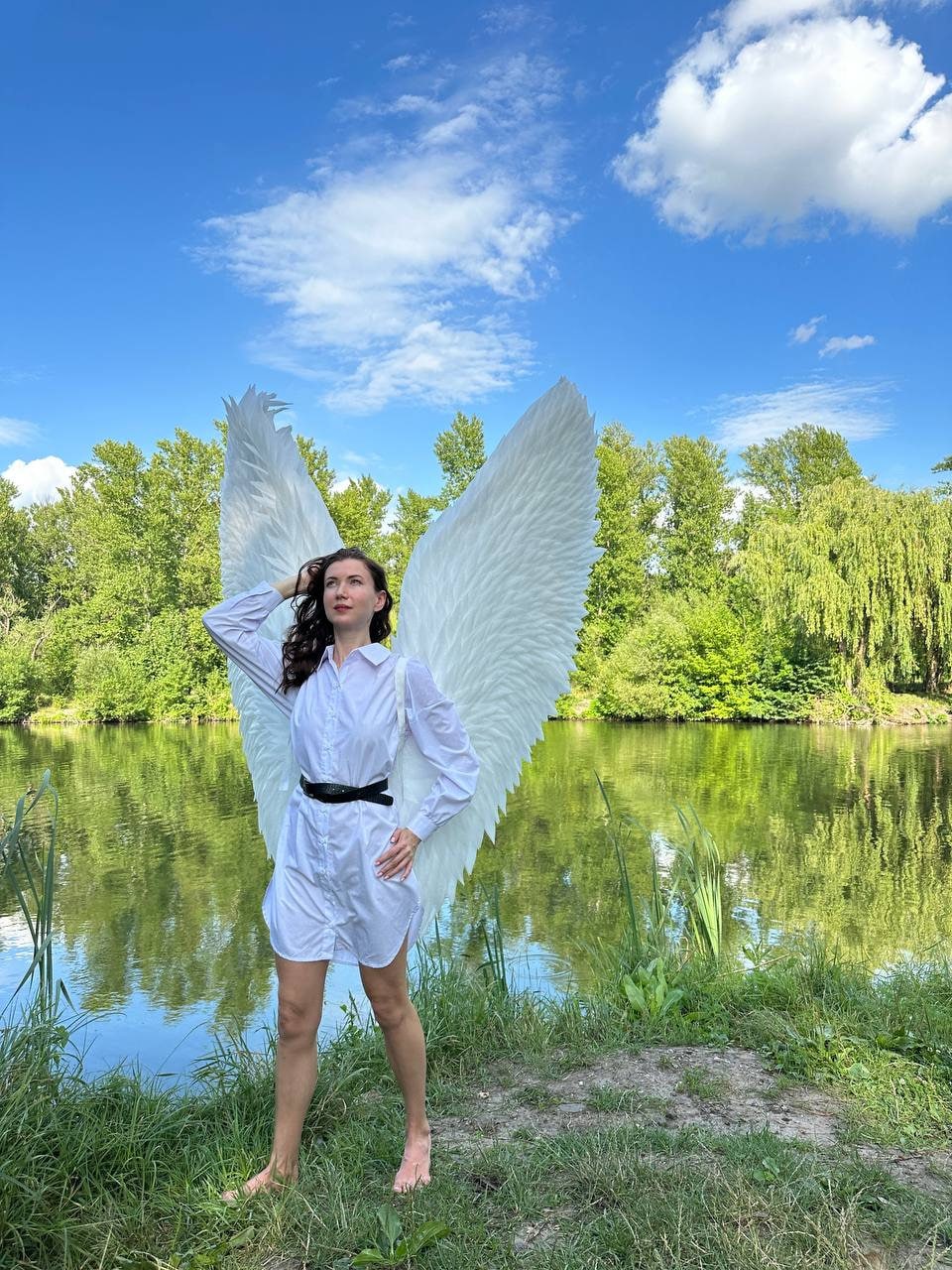 May include: A person wearing a white shirt and large white angel wings stands in a grassy area near a pond. The person is looking up at the sky.
