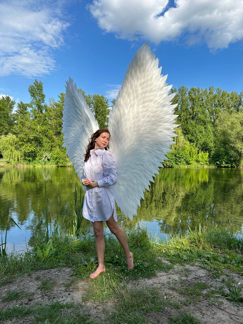 May include: A person wearing a white shirt stands by a lake with large white angel wings. The wings are made of feathers and are very detailed.