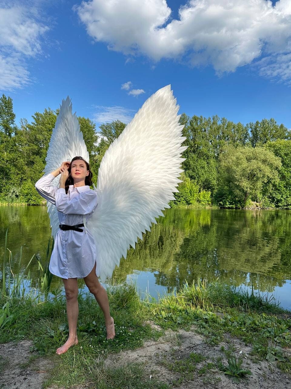 May include: A person wearing a white shirt and large white angel wings stands by a pond. The person is looking up at the sky.