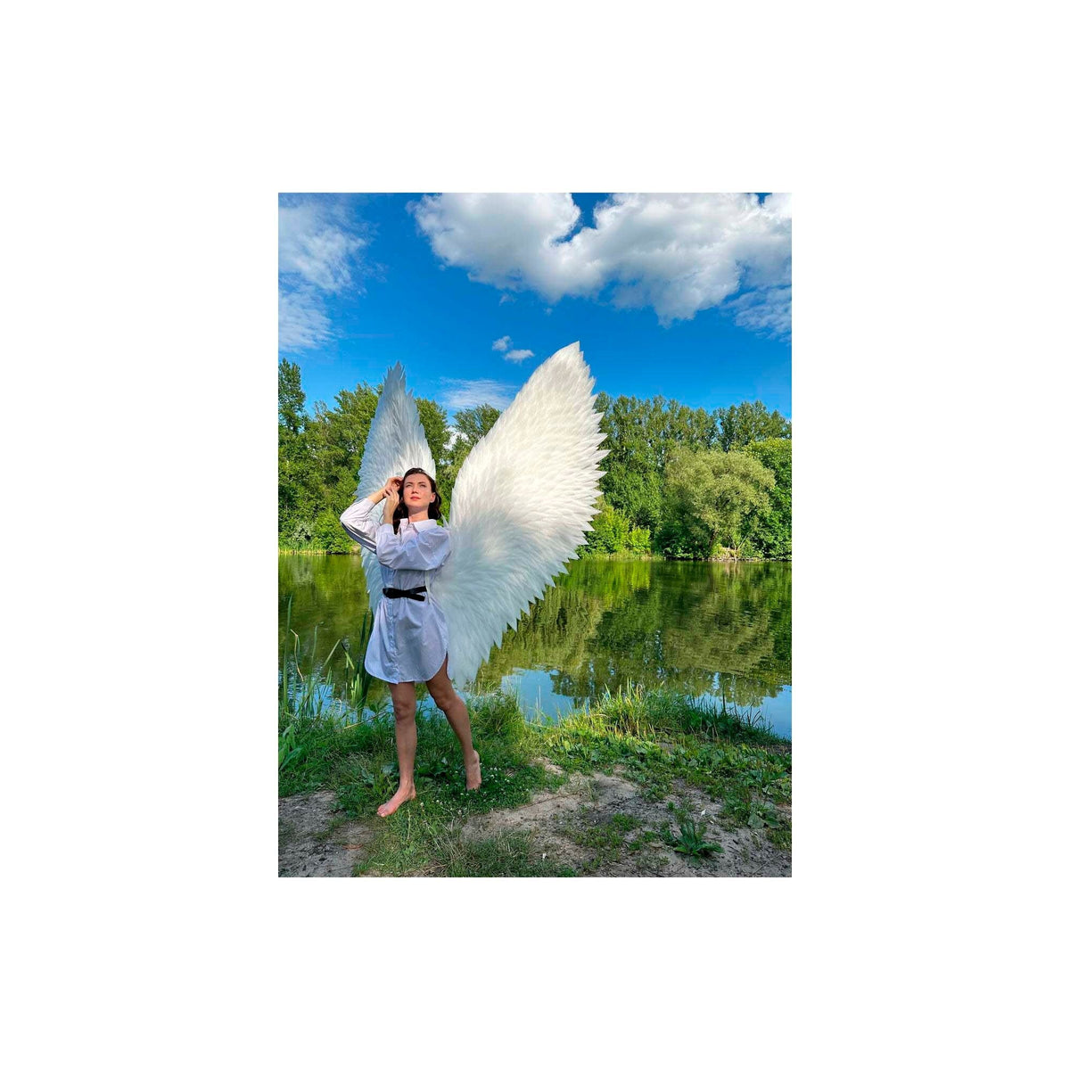 May include: A person wearing a white shirt and a black belt stands in front of a lake with large white angel wings. The wings are made of feathers and are spread out behind the person.