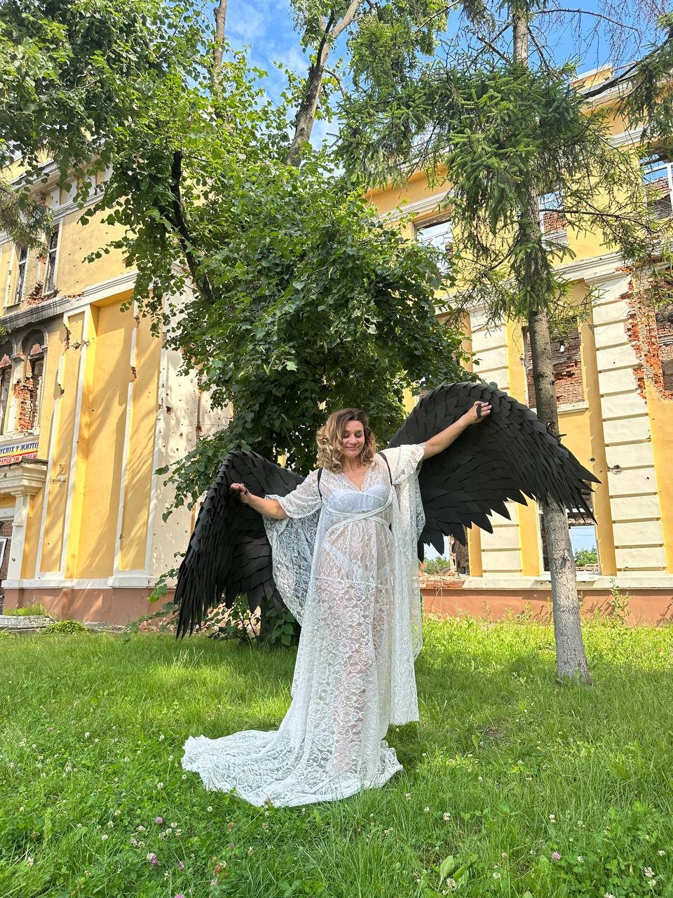 May include: A woman wearing a white lace gown and large black angel wings stands in a grassy area. The wings are spread wide, and the gown has a flowing train. The background includes a yellow building and trees.