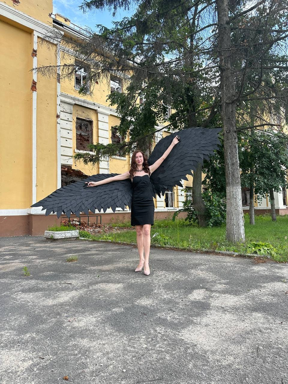 May include: A woman wearing a black dress and black feathered wings stands outdoors with arms outstretched. The wings are large and detailed, with each feather individually defined. The background includes a yellow building and trees.