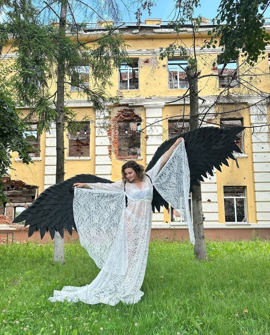 May include: A woman wearing a white lace dress and large black angel wings stands in front of a yellow building with visible damage. The wings are detailed and textured, contrasting with the delicate lace. The scene is set outdoors with green grass and trees.