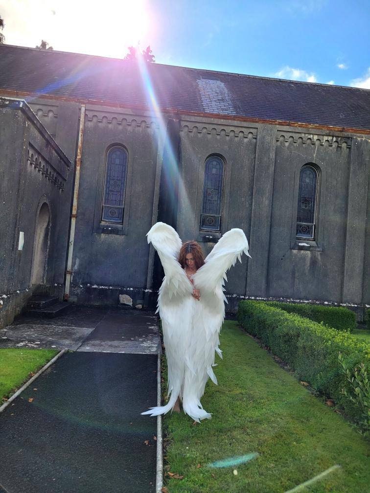 May include: A person wearing large, white feathered angel wings stands on a green lawn in front of a gray stone building. The wings are spread wide, and the person is facing the camera. The sky is blue with a bright sun.
