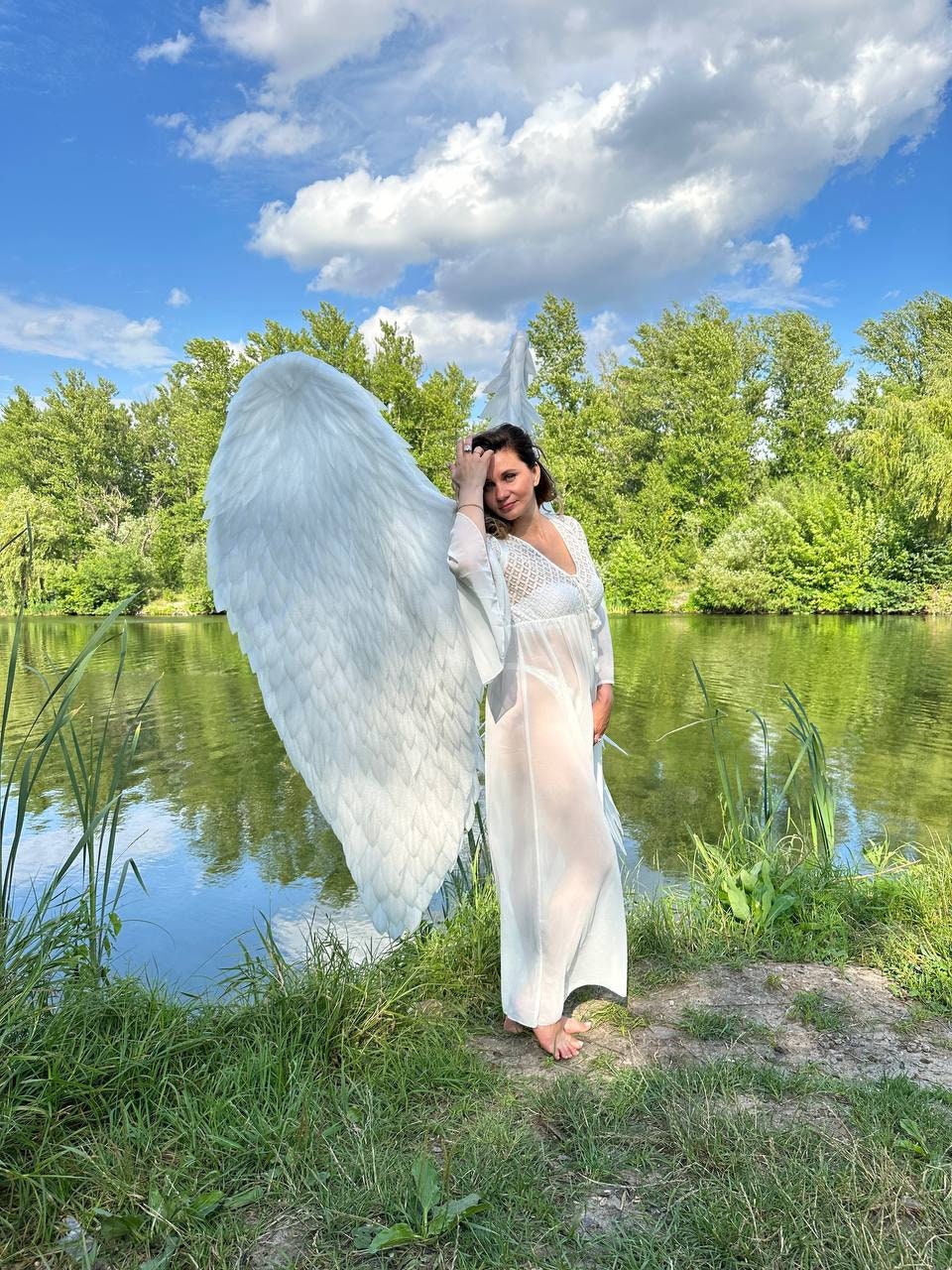 May include: A person wearing a white, flowing dress and large white feathered angel wings stands near a lake. The sky is blue with fluffy white clouds. The person is smiling and touching their hair.