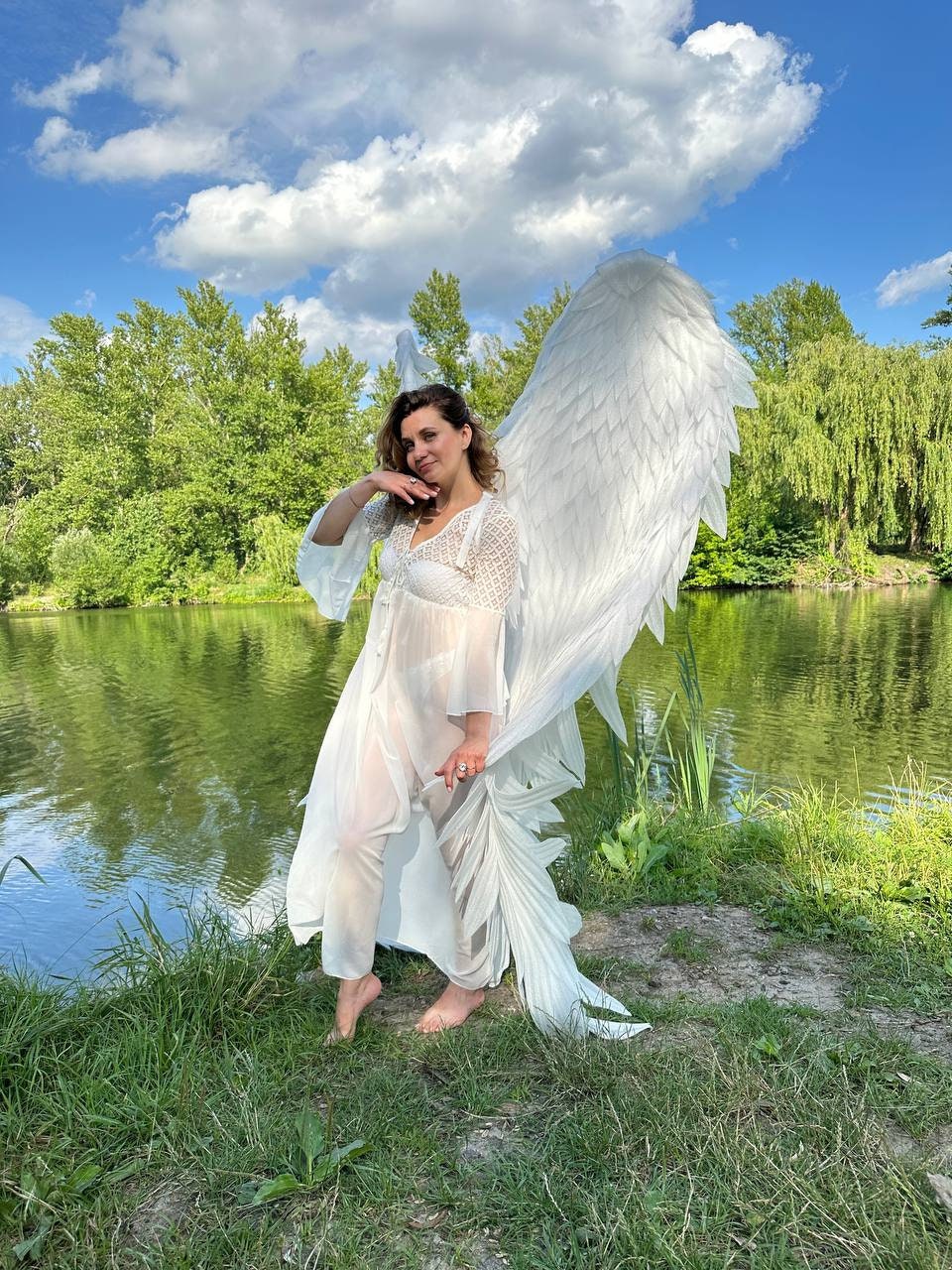 May include: A person wearing a white dress and large white angel wings stands near a lake. The wings are detailed with layered feathers. The sky is blue with fluffy white clouds, and green trees line the water's edge.