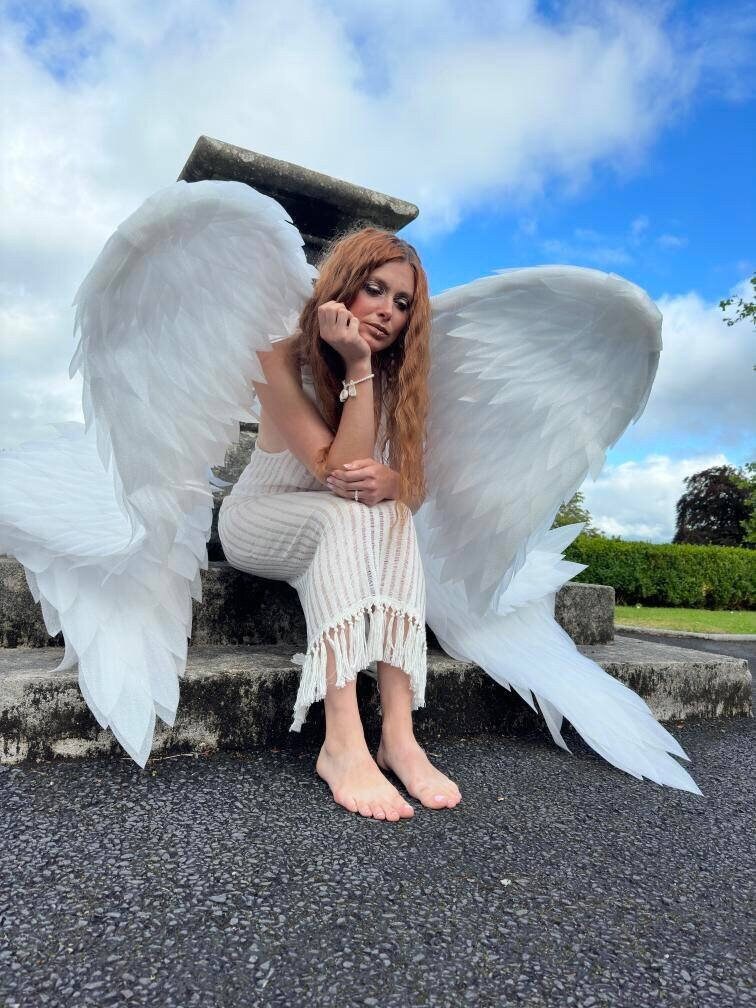 May include: A person wearing a white dress and large white angel wings sits on stone steps against a blue sky. The dress has a fringed hem. The person has long red hair and is wearing a bracelet.