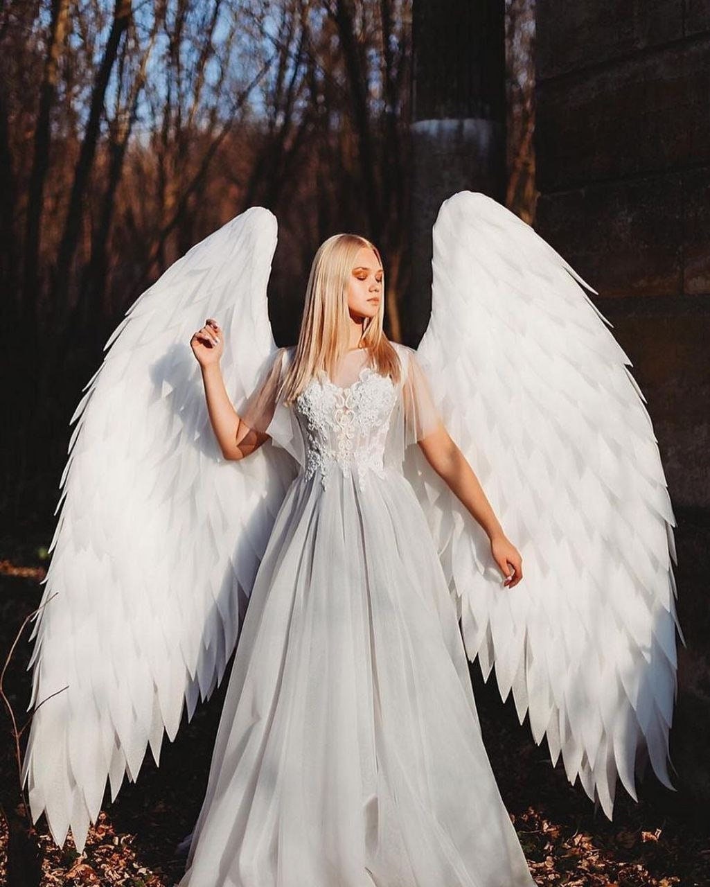 May include: A woman wearing a white dress with lace detailing and large white angel wings. The wings are made of feathers and are spread out behind her. She is standing in a forest setting.