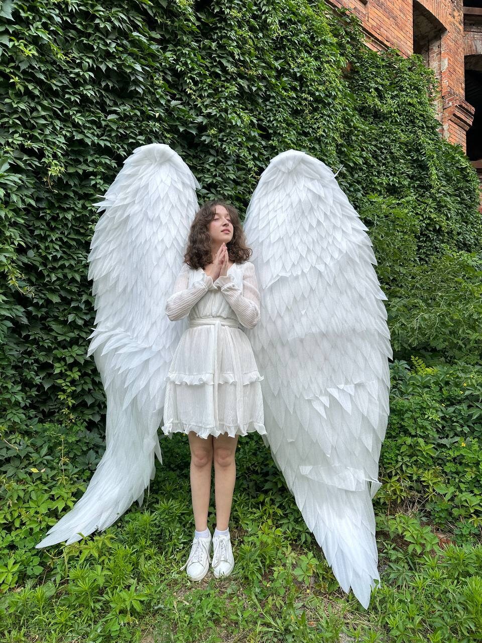 May include: A person wearing a white dress and white sneakers stands in front of a green wall with large white angel wings.