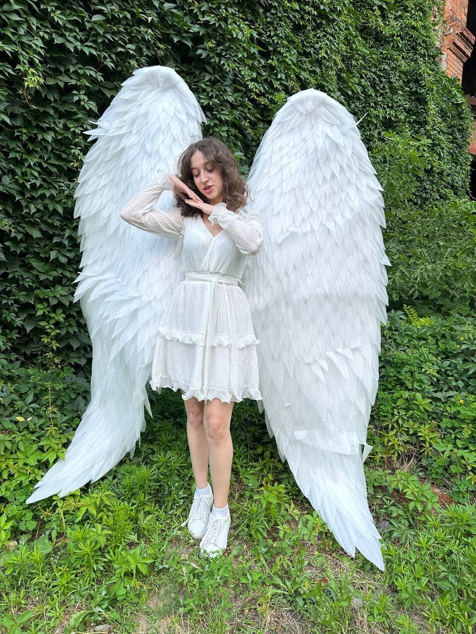 May include: A person wearing a white dress and white sneakers is standing in front of a green leafy background. They are wearing large white angel wings.
