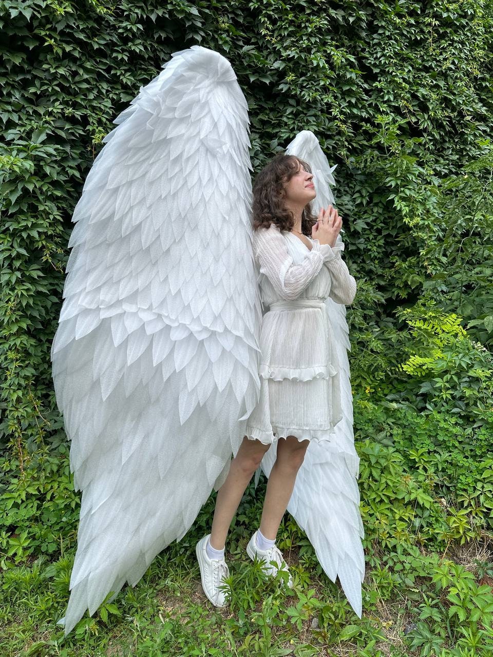 May include: A person wearing a white dress and white angel wings with a green leafy background.