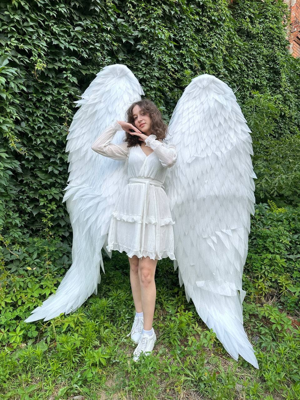 May include: A person wearing a white dress and white angel wings. The wings are large and feathery, and the person is standing in front of a green leafy background.