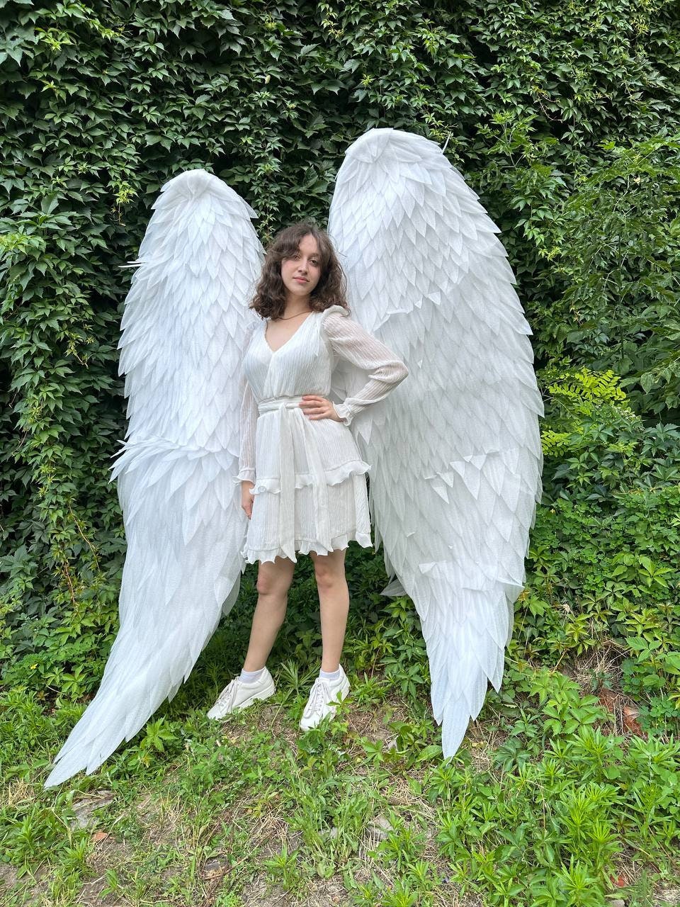 May include: A person wearing a white dress and white sneakers is standing in front of a green leafy background. They are holding large white angel wings.