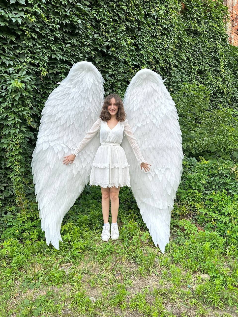 May include: A person wearing a white dress and white sneakers stands in front of a green wall with large white angel wings.