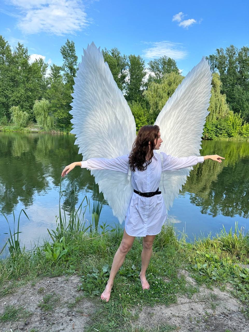 May include: A person wearing a white shirt and large white angel wings stands by a lake. The wings are made of feathers and are very detailed. The person is looking to the right and has long brown hair.