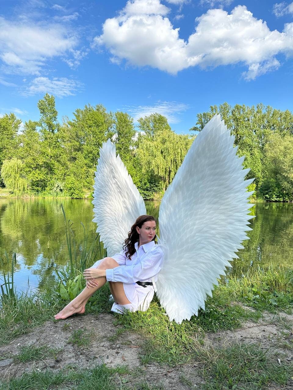 May include: A person wearing a white shirt and large white angel wings sits by a pond with green grass and trees in the background.