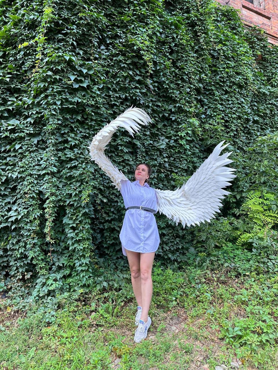 May include: A person wearing a light blue shirt and white sneakers is standing in front of a green ivy wall. They are holding large white wings made of paper or feathers.