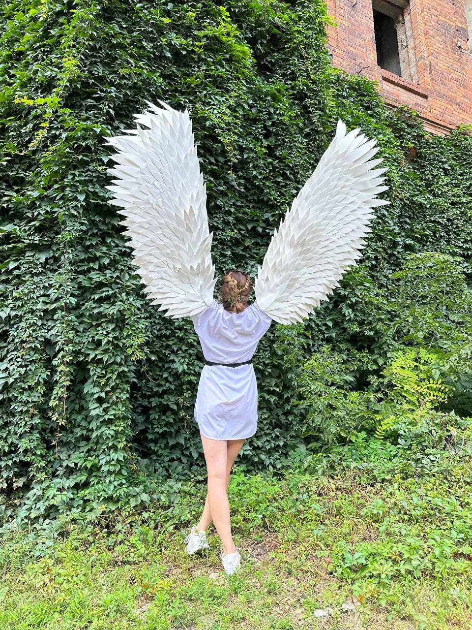 May include: A person wearing a white shirt and white sneakers with large white angel wings. The person is standing in front of a green ivy covered wall.