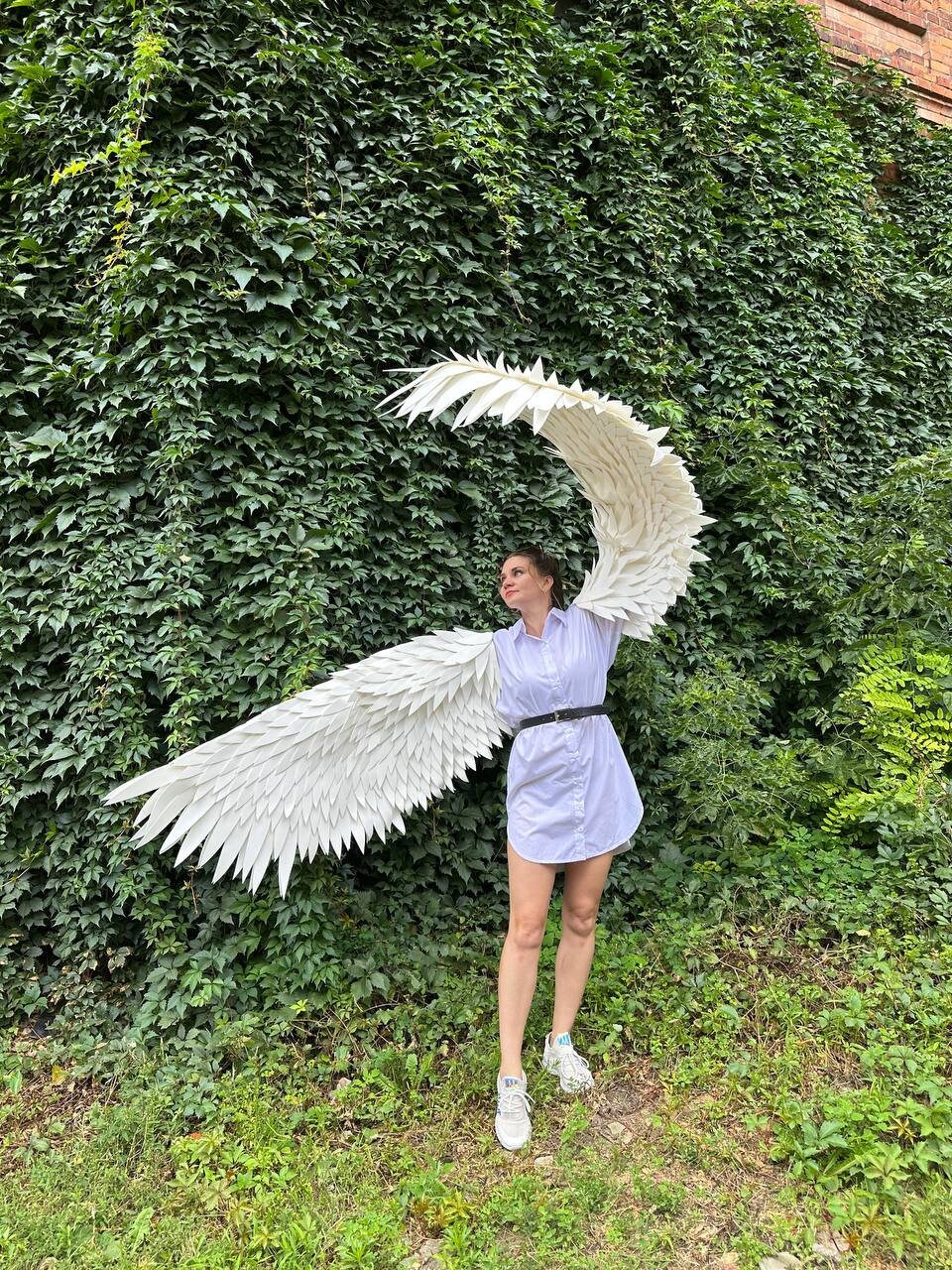 May include: A person wearing a white shirt and jeans is holding large white angel wings in front of a green ivy wall.