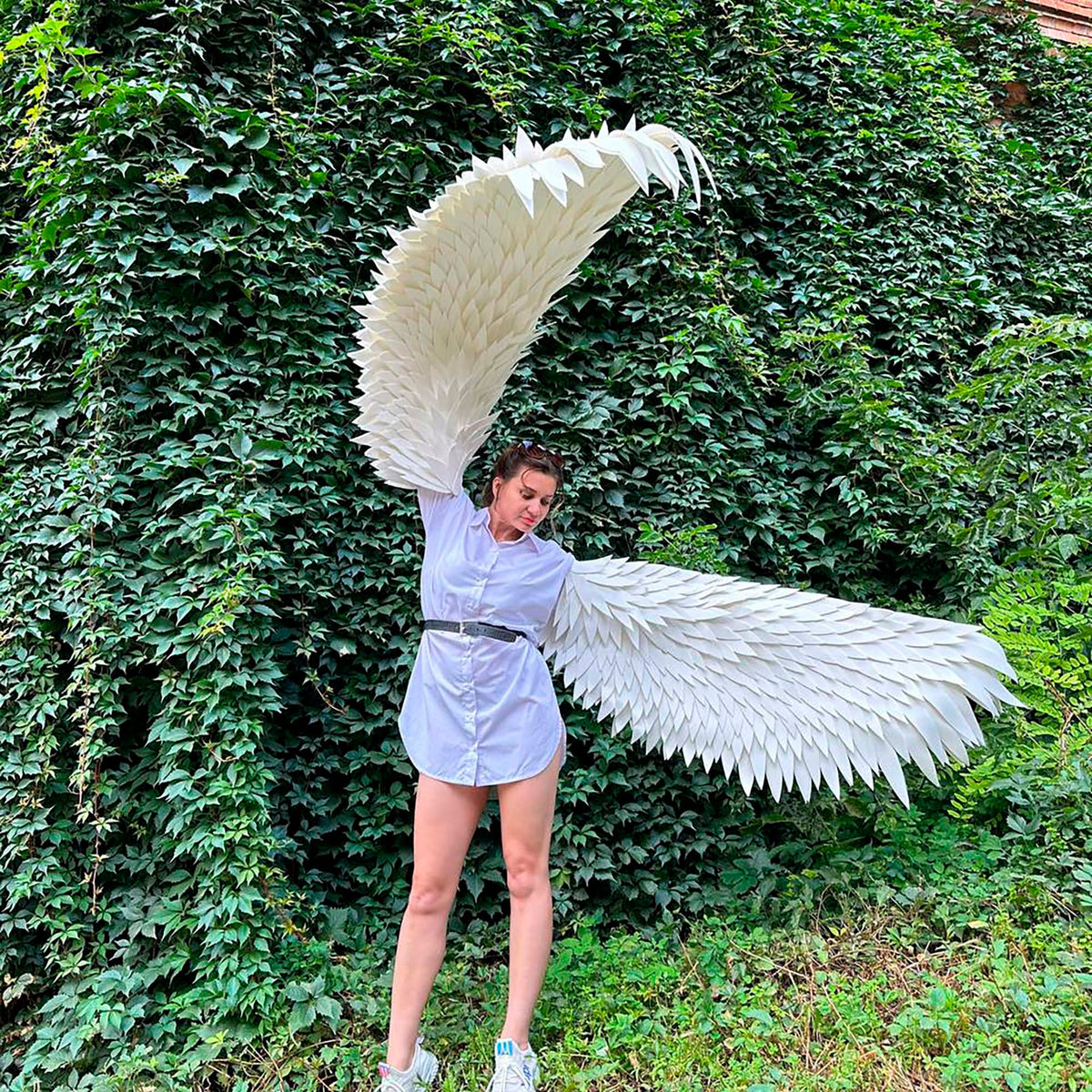 May include: A person wearing a white shirt and a black belt is holding large white angel wings in front of a green ivy wall.