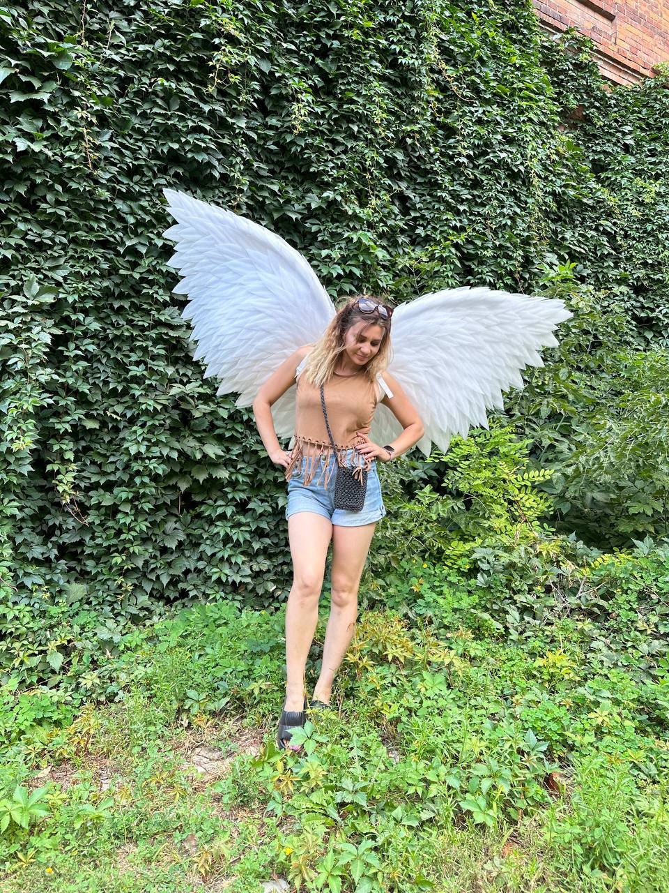 May include: A woman wearing a brown tank top and denim shorts is standing in front of a green wall. She is wearing a pair of white angel wings.