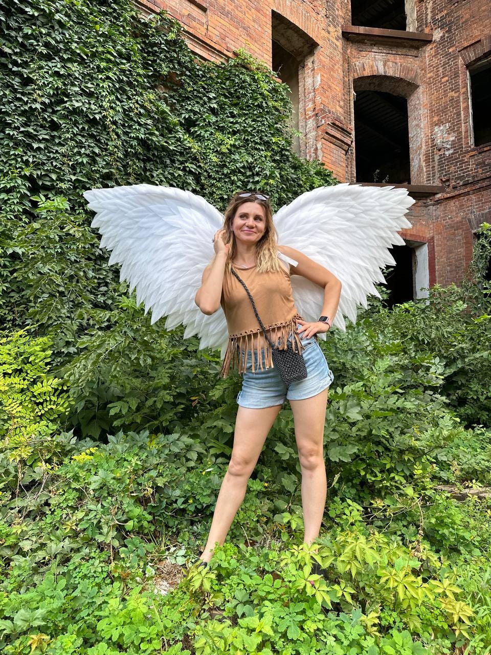 May include: A woman wearing a brown tank top with fringe, denim shorts, and white angel wings. She is standing in front of a brick building covered in green vines.