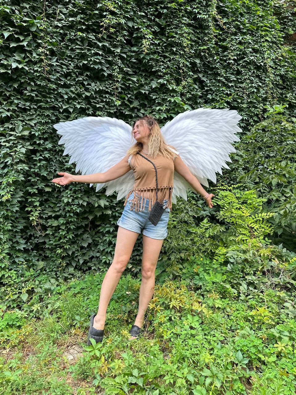 May include: A person wearing a brown tank top and denim shorts is standing in front of a green leafy wall. They are holding large white angel wings out to the sides.