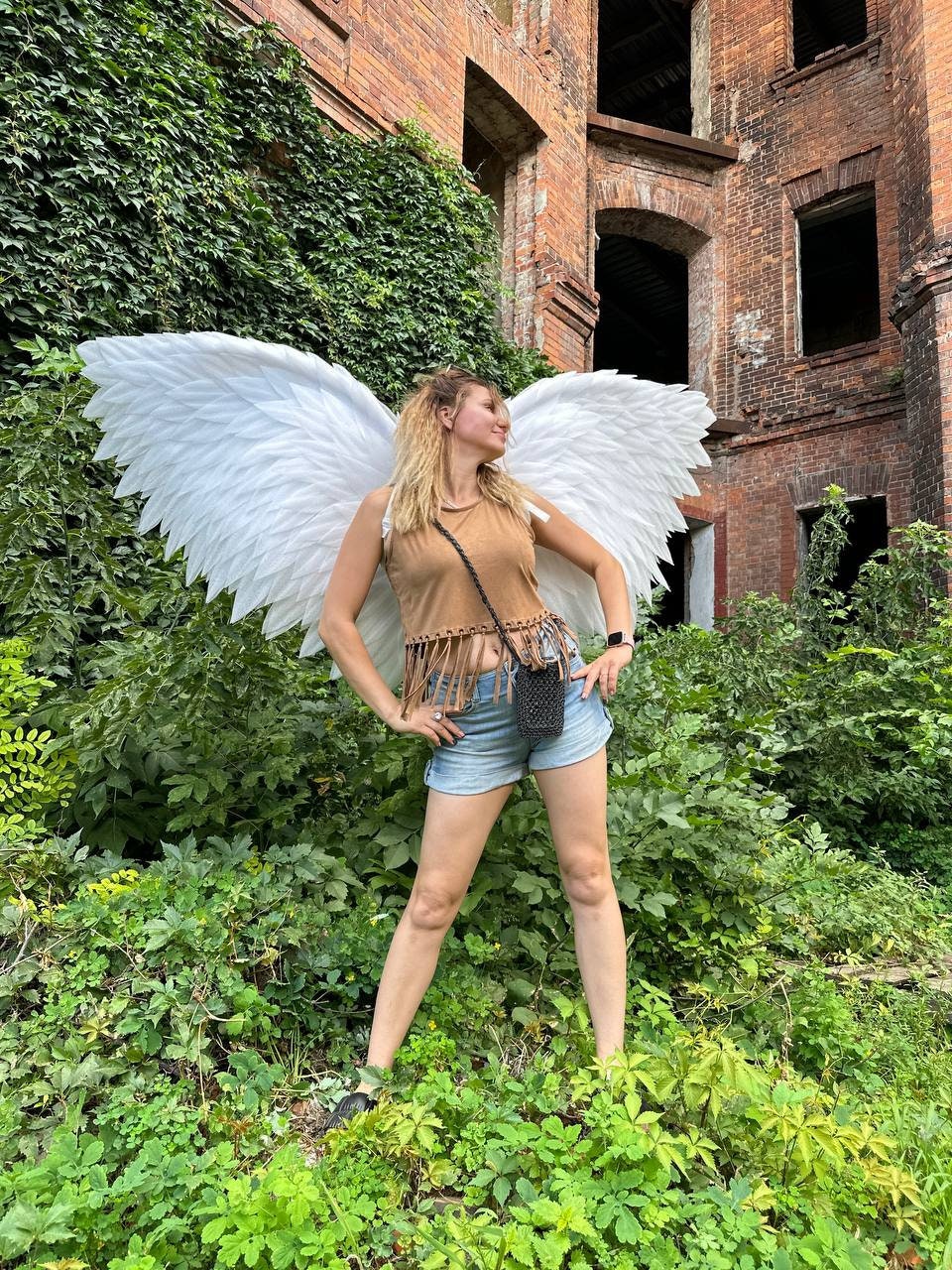 May include: A woman wearing a brown tank top and denim shorts stands in front of a brick building with ivy growing on the walls. She is wearing large white angel wings.