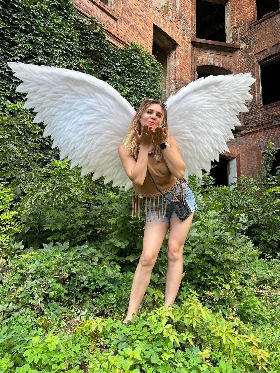 May include: A woman wearing a brown top and denim shorts with white angel wings. She is standing in front of a brick wall covered in green vines.