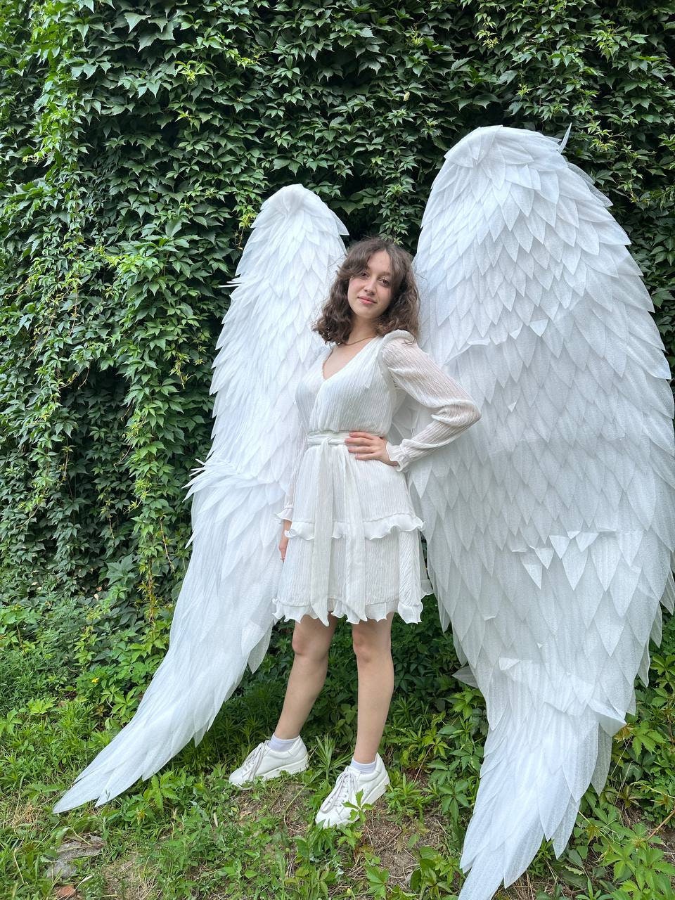 May include: A person wearing a white dress and white sneakers stands in front of a green leafy wall with large white angel wings.