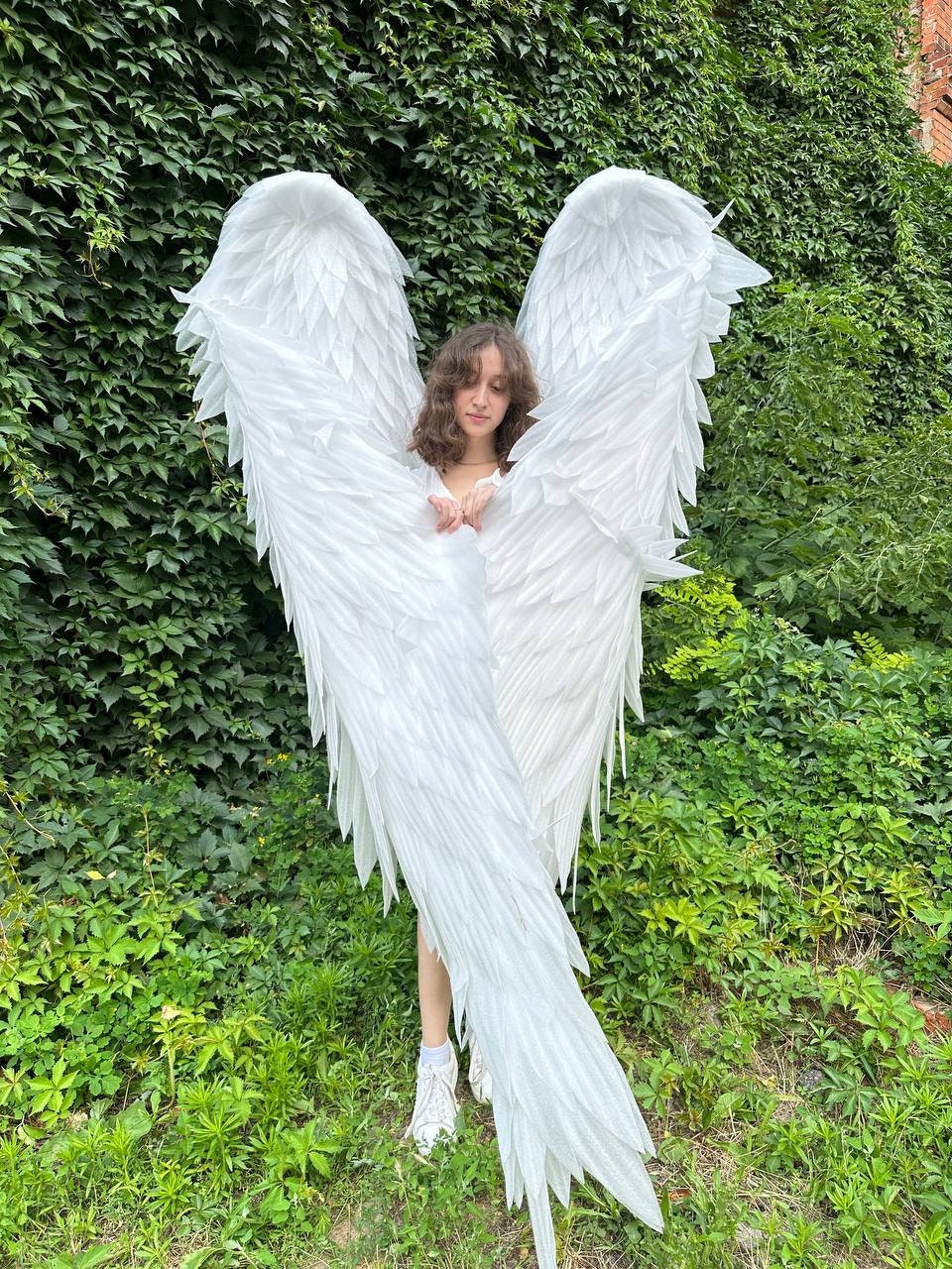 May include: A person wearing a white dress and white angel wings with a green leafy background.