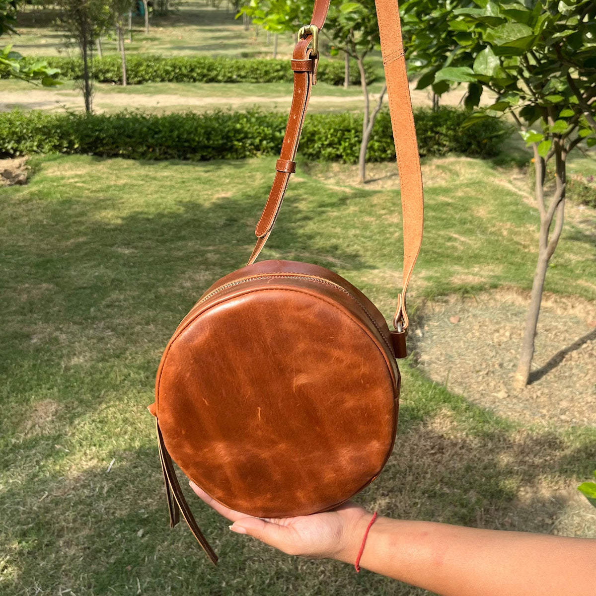 May include: A round brown leather crossbody bag with a zipper closure and a long strap. The bag has a fringe detail on the bottom.