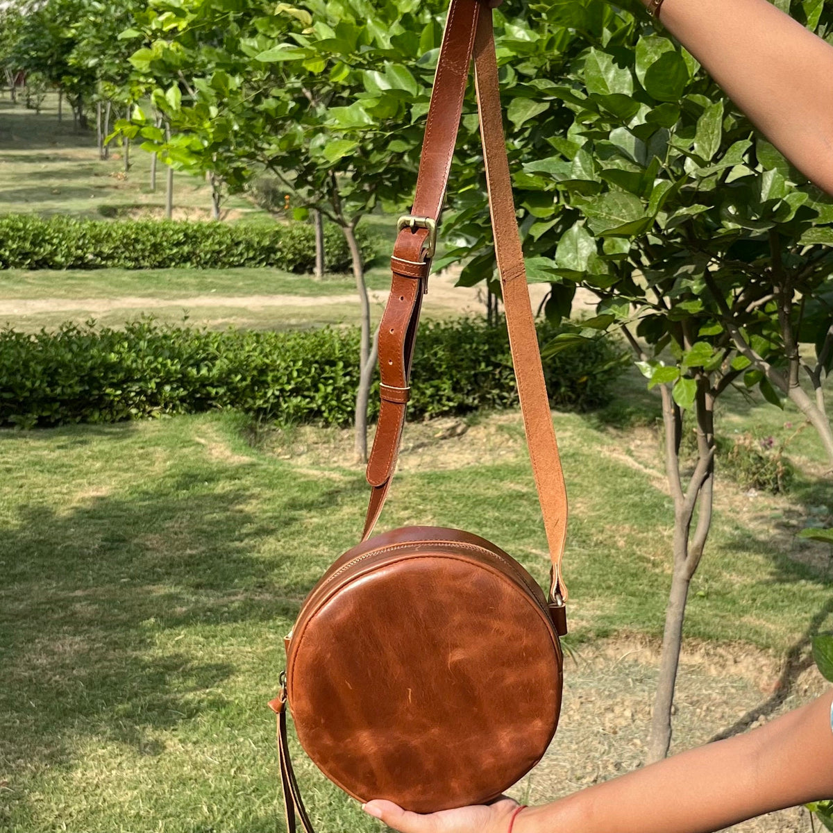 May include: A brown leather round crossbody bag with a long adjustable strap. The bag has a flap closure and a tassel.